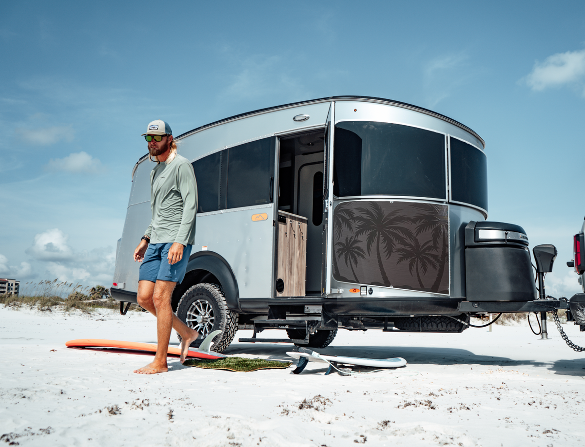 A man in a hat and sunglasses stands barefoot on sand near surfboards, next to a modern silver Basecamp X camper trailer featuring the Airstream Rock Guard Rock Wraps Nature Collection with palm tree graphics, under a blue sky.
