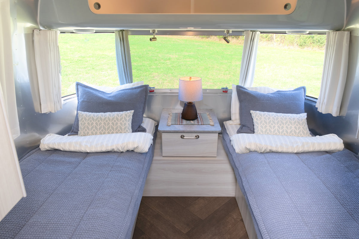 Airstream Custom Fit Dual Comfort Beddy’s for Globetrotter Travel Trailers on two twin beds in an Airstream, each featuring a blue quilted comforter, two pillows, and a small nightstand between them.