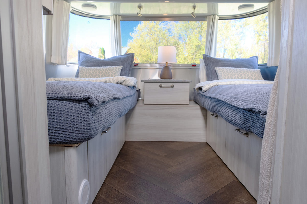 Two twin beds in an Airstream featuring Airstream Custom Fit Dual Comfort Beddy’s for Trade Wind Travel Trailers, both made with textured blue bedding and accent pillows, separated by a nightstand with a lamp before a panoramic window.