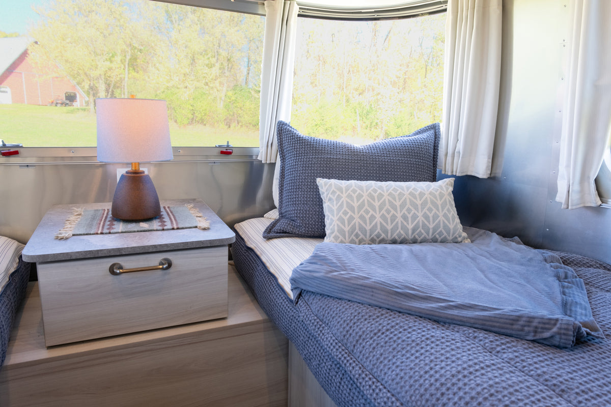 A twin-sized bed made with a blue Airstream Custom Fit Dual Comfort Beddy’s Flying Cloud Travel Trailers bedding system, a patterned throw pillow, and a bedside table with a lamp, set against a curved wall with a window.