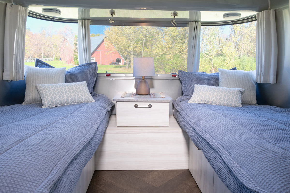Two twin beds featuring Airstream Custom Fit Dual Comfort Beddy’s Flying Cloud Travel Trailers with blue quilted covers and accent pillows, separated by a nightstand with a lamp inside a travel trailer looking out onto a rural landscape.