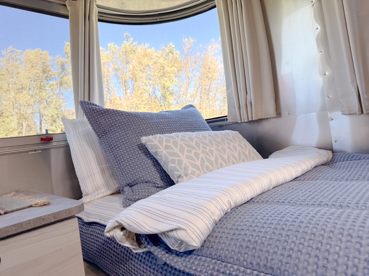 A bed in a travel trailer with curved windows, featuring an Airstream Custom Fit Dual Comfort Beddy’s for Globetrotter Travel Trailers with a quilted blue cover and patterned pillows.