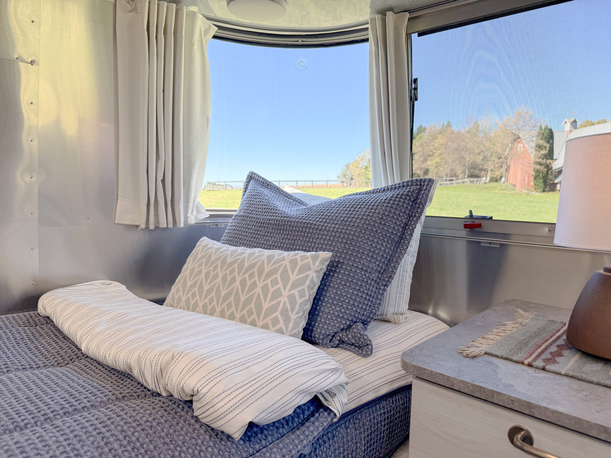 A made bed featuring a blue waffle-knit Airstream Custom Fit Dual Comfort Beddy’s for Globetrotter Travel Trailers sits in a curved Airstream bedroom next to a window overlooking a grassy field.