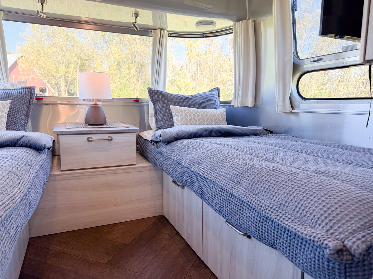 Two twin beds in a travel trailer outfitted with Airstream Custom Fit Dual Comfort Beddy’s for Stetson + 6666 Travel Trailers, each featuring a Dual Comfort ZipBase and dark blue textured bedding with coordinating pillows.