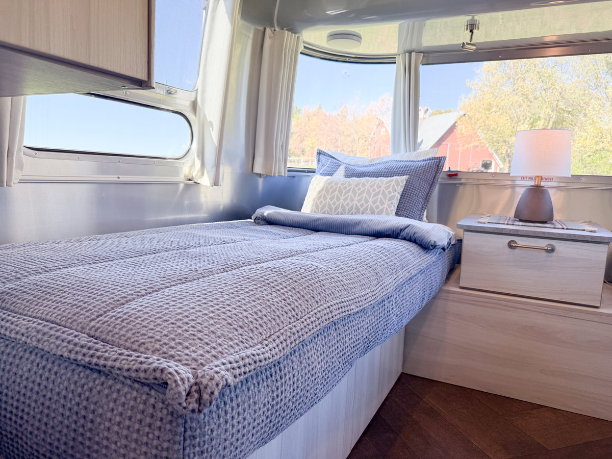 A twin-size Airstream Custom Fit Dual Comfort Beddy’s for Pottery Barn Travel Trailers comforter in a blue textured fabric and a patterned accent pillow styled on a bed inside a travel trailer.
