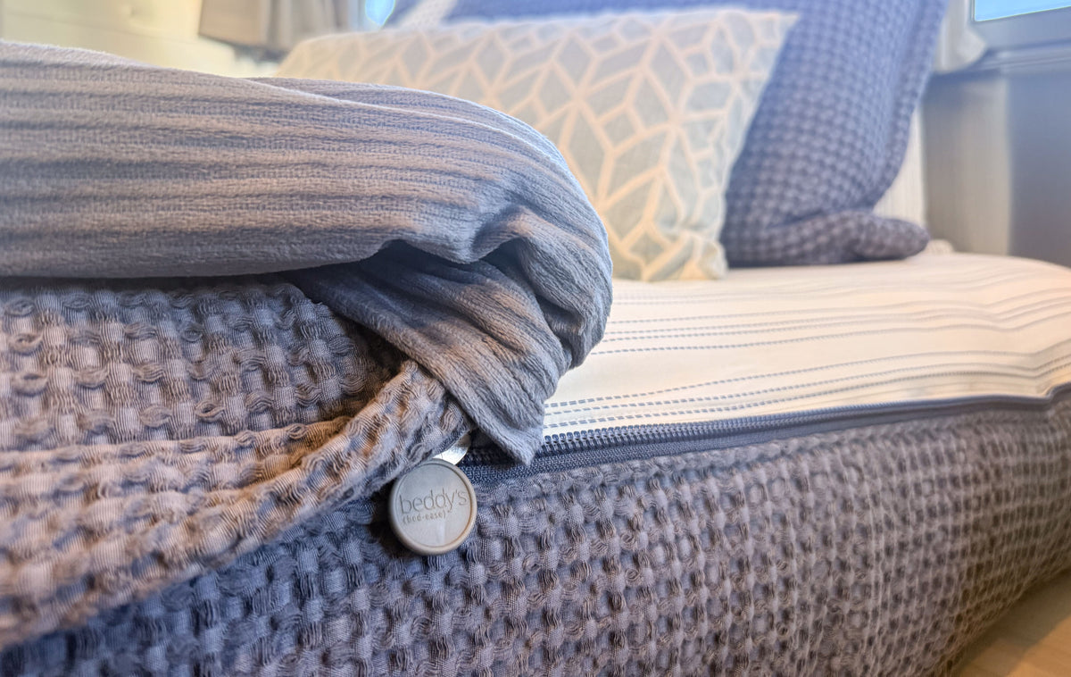 A gray zippered Airstream Custom Fit Dual Comfort Beddy’s for Pottery Barn Travel Trailers bedding set on a mattress, featuring a circular logo tag on the side and patterned decorative pillows in the background.