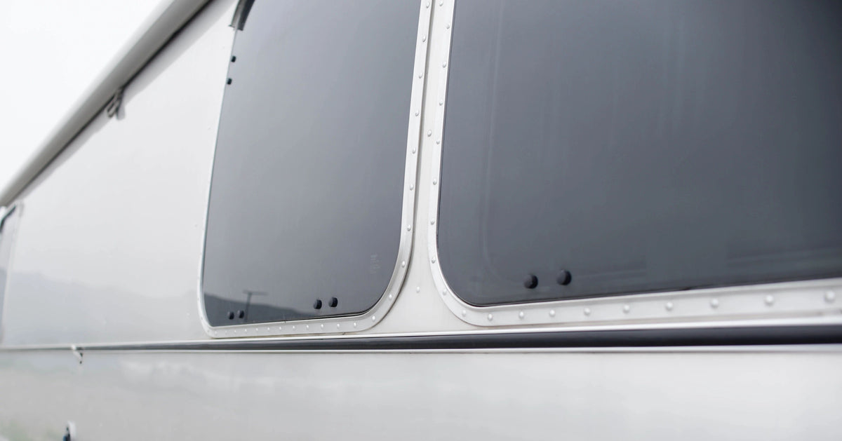 Close-up of two large, tinted, rectangular windows with rounded edges and visible rivets on a sleek, silver metal surface—possibly part of a travel trailer or RV—featuring the Airstream Premium Replacement Belt Trim for Travel Trailers as an essential exterior accessory.