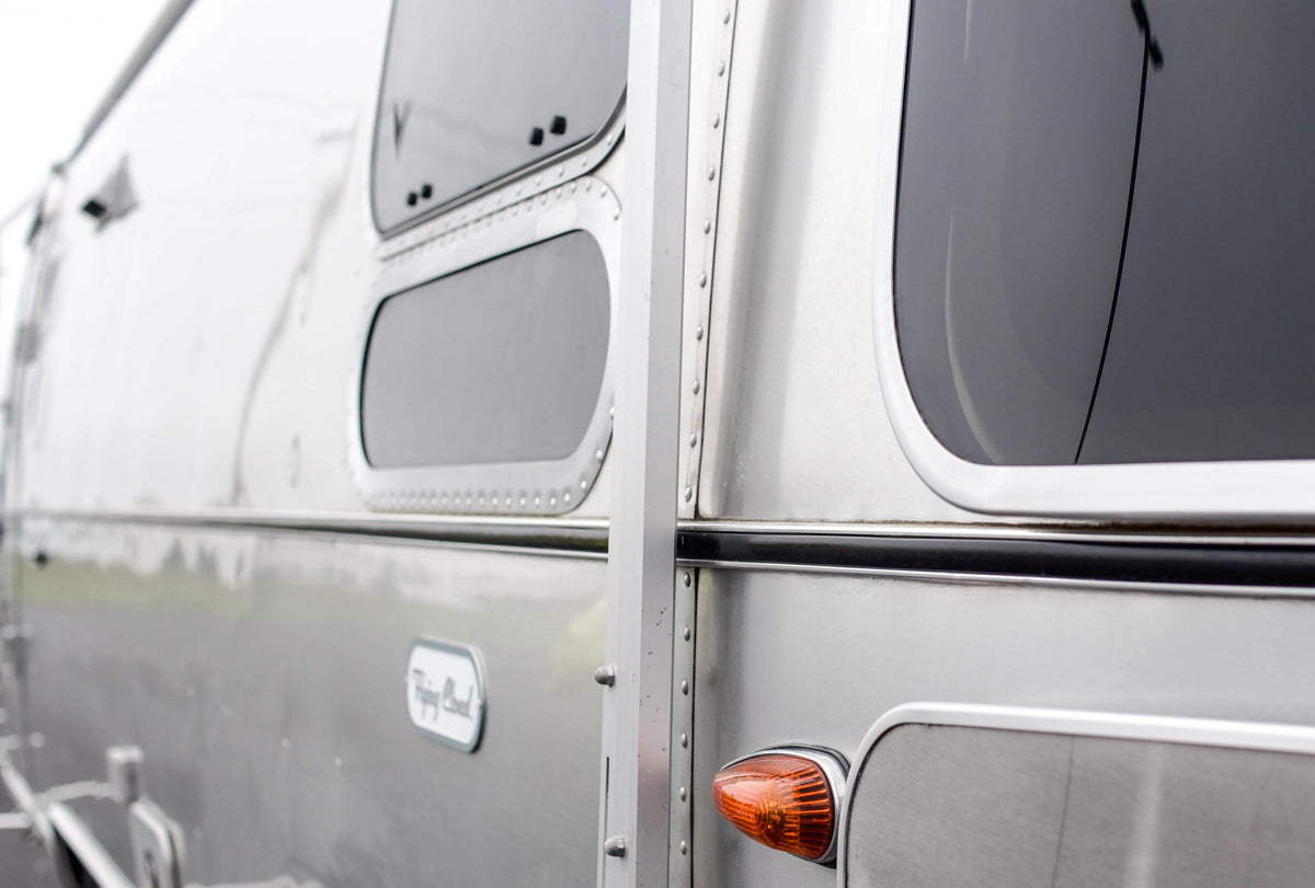 Close-up of the side of a shiny silver Airstream trailer, highlighting windows, rivets, an orange side light, and part of the logo near the bottom. The expertly crafted Airstream Premium Replacement Belt Trim for Travel Trailers is clearly visible along the edge. The background is blurred.