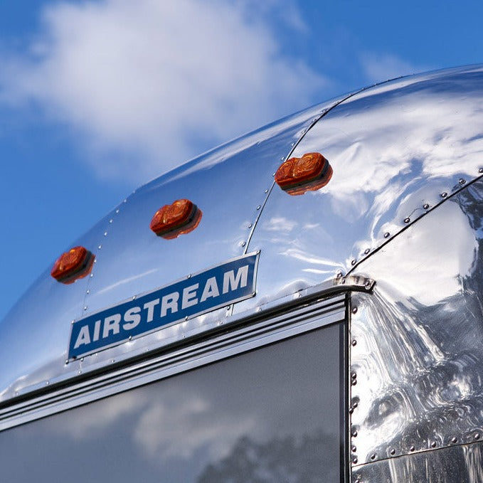 Close-up of a shiny silver Airstream travel trailer with an authentic Airstream 1960′s Aluminum Blue Logo Nameplate (Part #51101WR) and orange lights on its curved top, reflecting the blue sky and clouds above.