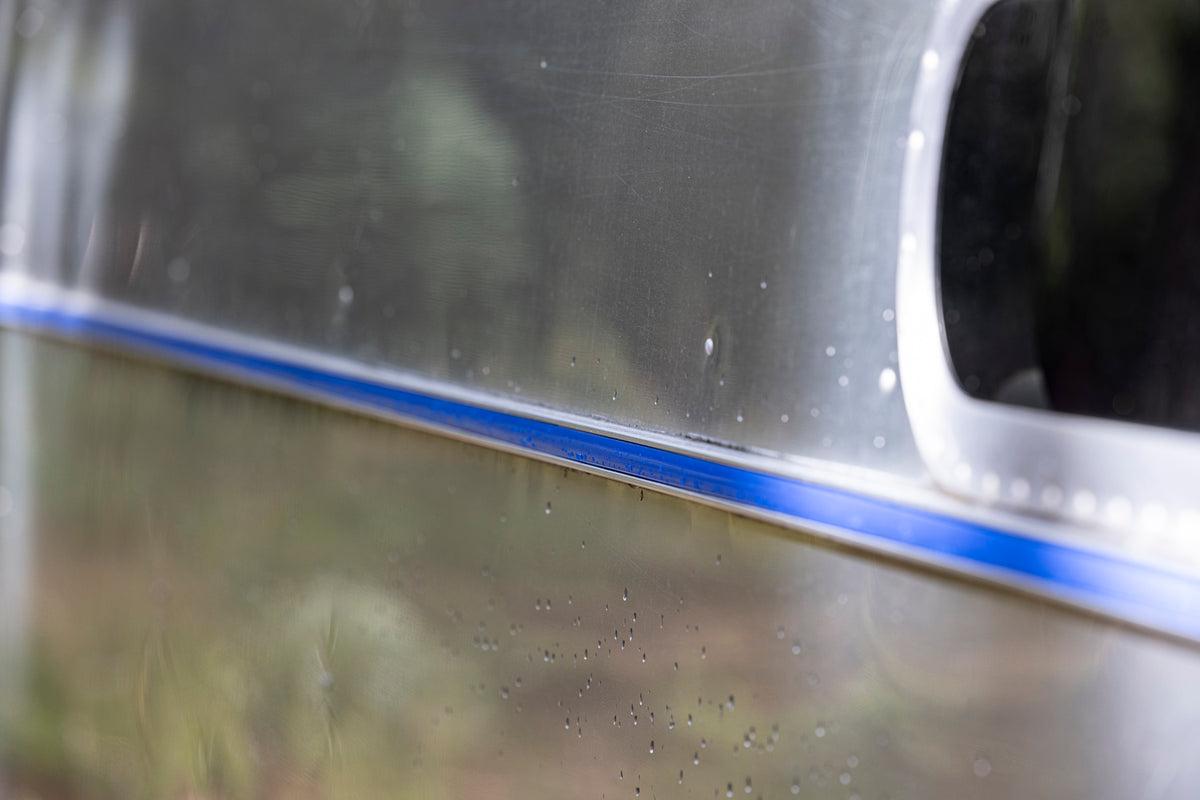 Close-up of the Airstream Premium Replacement Belt Trim for Travel Trailers, showcasing its shiny metallic aluminum surface with a blue horizontal stripe, water droplets, and a rounded window on the right side; blurred greenery in the background.