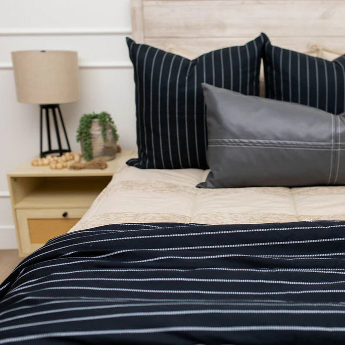 A neatly made bed features the Bryant Luxe Blanket by Beddy’s, showcasing navy blue and gray stripes, complemented by two large pillows and a gray accent pillow. Beside the bed sits a nightstand with a lamp and a potted plant.