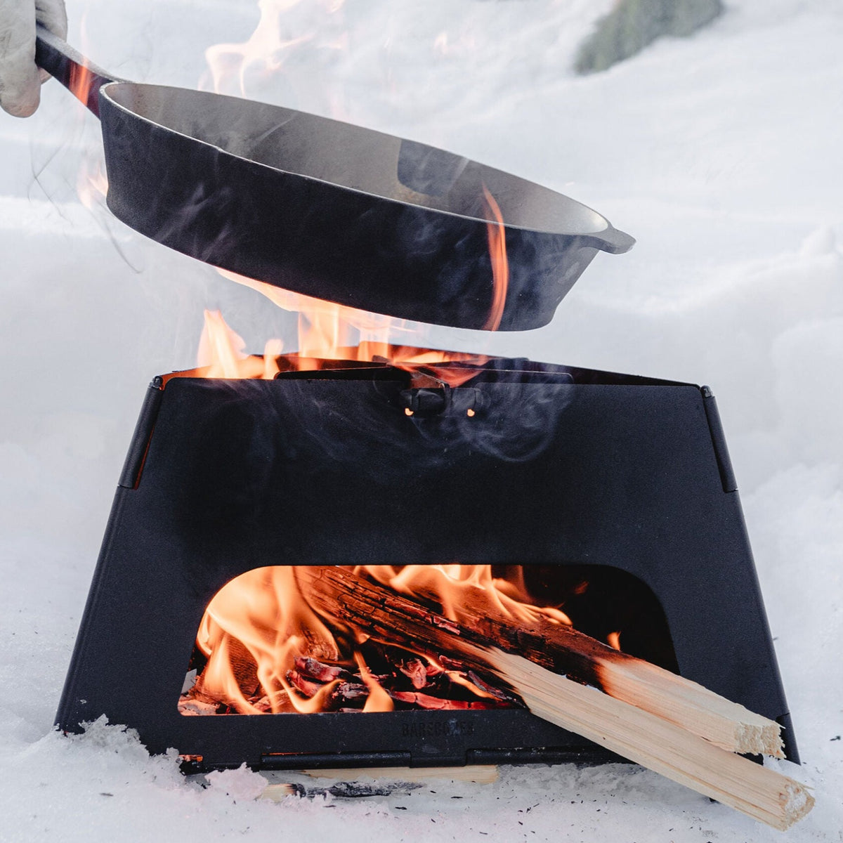 The Flatbrook Folding Stove by Barebones, filled with burning wood, is being used to support a cast iron pan in a snowy landscape.