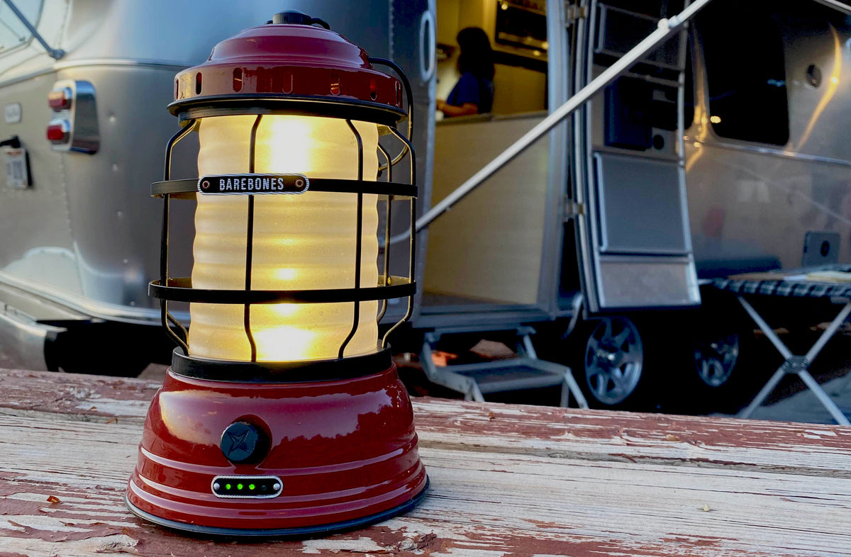 A red Barebones lantern glows on a weathered picnic table, with a silver camper and steps visible in the blurred background.