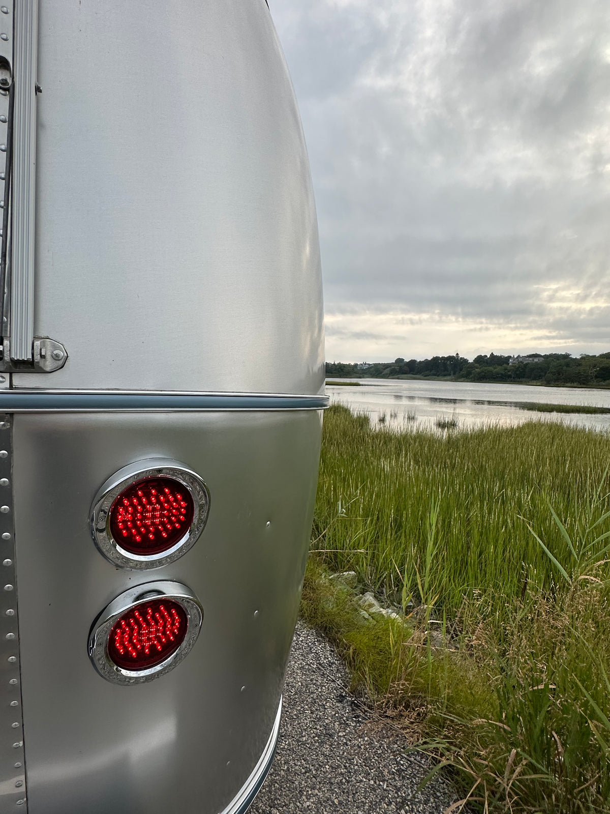 Close-up of a silver trailer’s rear lights, glowing red, parked beside a grassy marshland under a cloudy sky. The sleek Airstream Premium Replacement Belt Trim for Travel Trailers accents the exterior, highlighting the trailer’s iconic aluminum design.