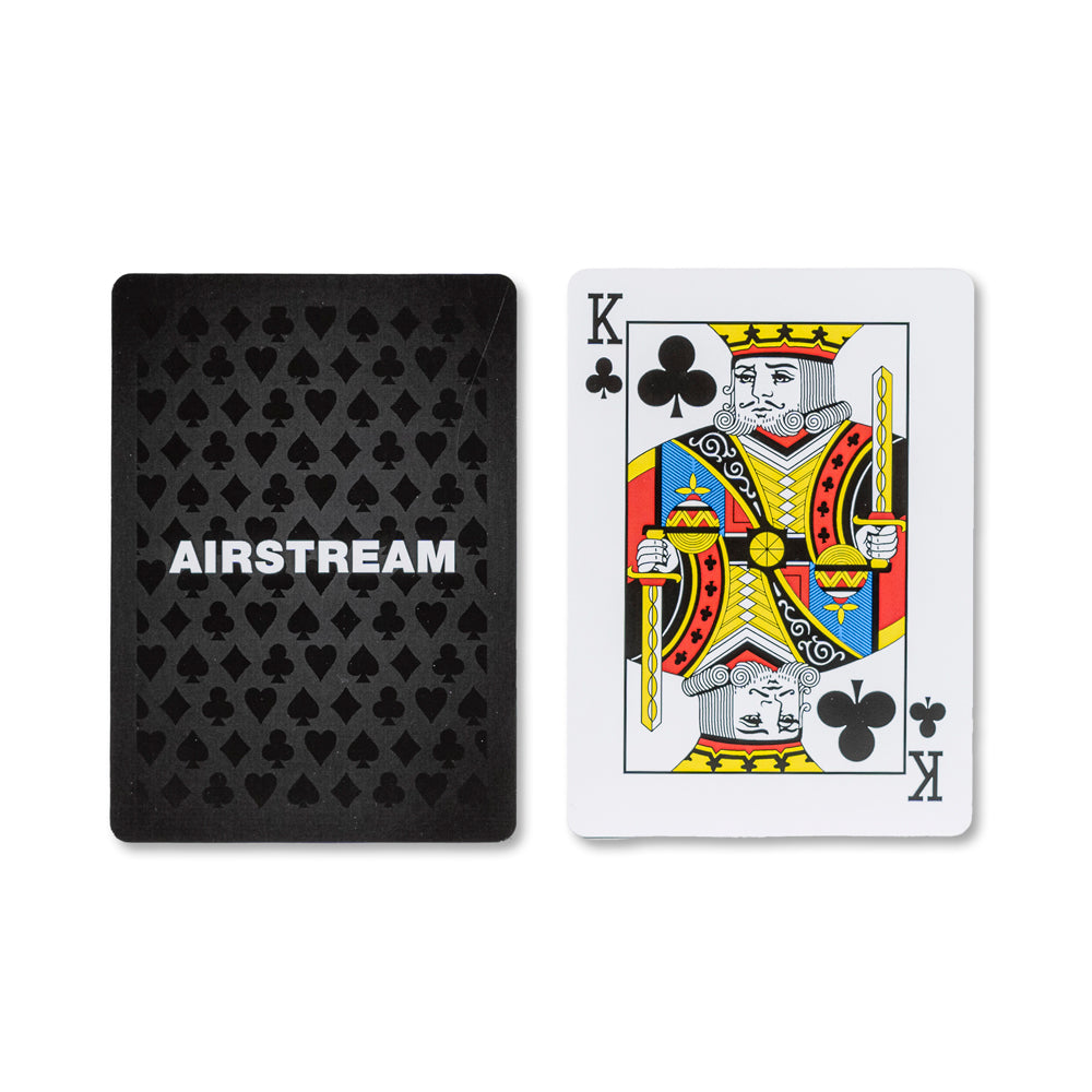 Two playing cards from the Airstream Card Deck With Case are shown: the back of one card features a black design with “AIRSTREAM” and spade symbols, while the front displays a traditional King of Clubs in black, yellow, red, and blue. This stylish travel card game is part of a premium deck with case—perfect for fun on the go.