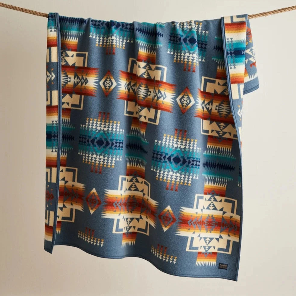 The Chief Joseph Blanket Robe by Pendleton, featuring geometric patterns in blue, orange, cream, and green, hangs over a rope against a plain wall—a design inspired by Native American gifting traditions.