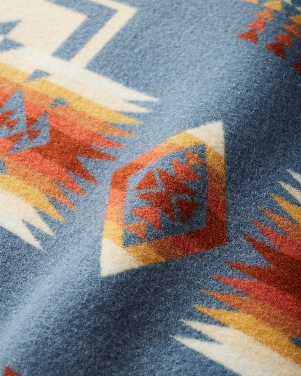 Close-up of the Chief Joseph Blanket Robe by Pendleton, showcasing its soft wool fabric with a blue base and geometric patterns in red, gold, white, and orange. Inspired by traditional Native American designs, the blanket’s surface appears textured and slightly fuzzy.