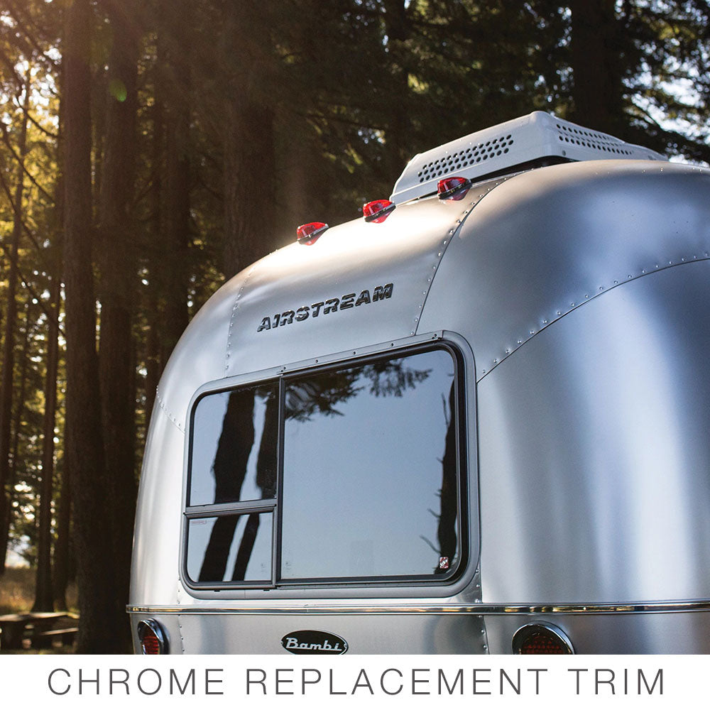 A close-up of the rear of an Airstream trailer featuring the Airstream Premium Replacement Belt Trim for Travel Trailers, with polished aluminum and chrome detailing reflecting the surrounding trees and sky, parked among tall trees. Text at the bottom reads CHROME REPLACEMENT TRIM.