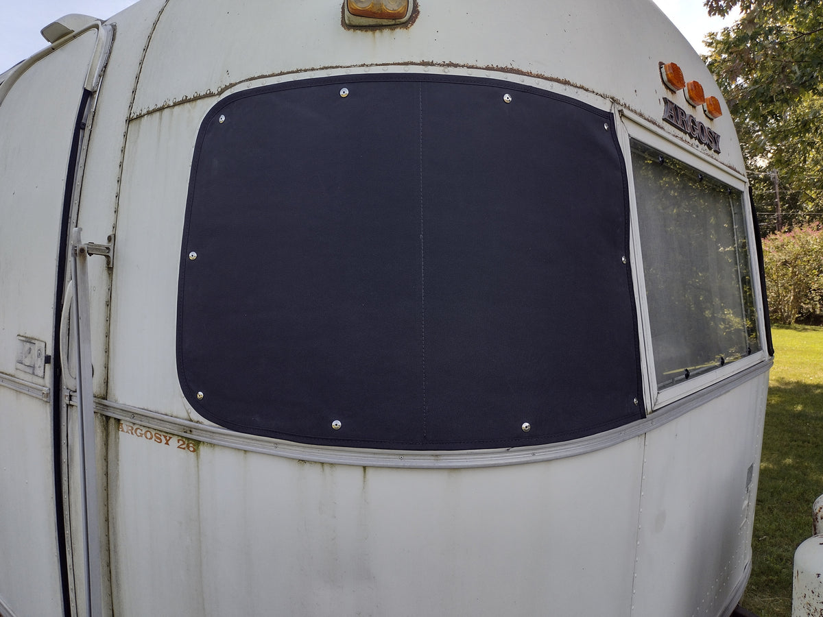 A close-up of an old white trailer features the Airstream Padded Window Protector for Argosy, a dark, rectangular cover securely fastened over the window. The trailer displays signs of rust and weathering, with grass and trees in the background.