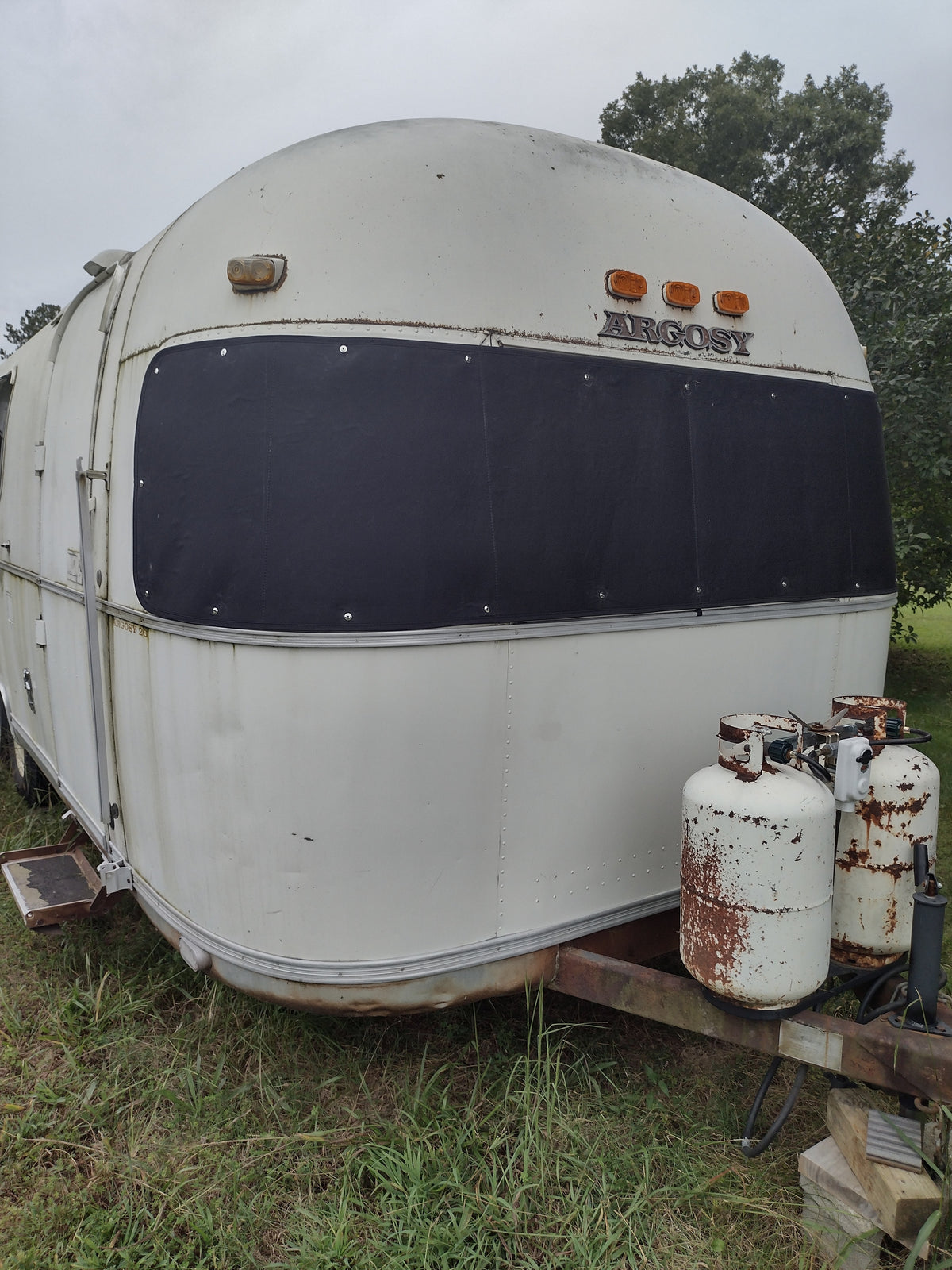 A vintage white Argosy trailer sits on grass, with two rusty propane tanks attached to the front hitch and an Airstream Padded Window Protector for Argosy shielding its panoramic window. Trees and a cloudy sky are visible in the background, completing this timeless scene accented by classic vintage travel trailer accessories.
