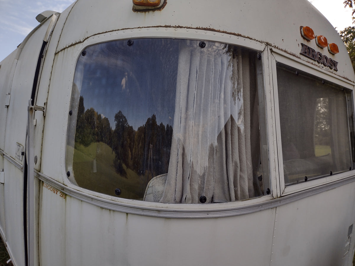 Close-up of an old white camper with dirty, partially torn curtains and trees reflected in the large front window under a cloudy sky. The Airstream Padded Window Protector for Argosy, one of several vintage travel trailer accessories, adds both protection and character to this classic vehicle.