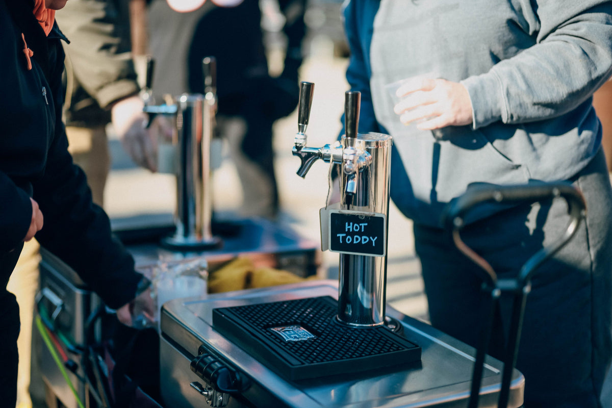A person stands near The Cooler Keg, a portable draft system, at an outdoor event, holding a clear cup. Another attendee is reaching for something on the table, enjoying the convenience of outdoor beverages served from The Cooler Keg.