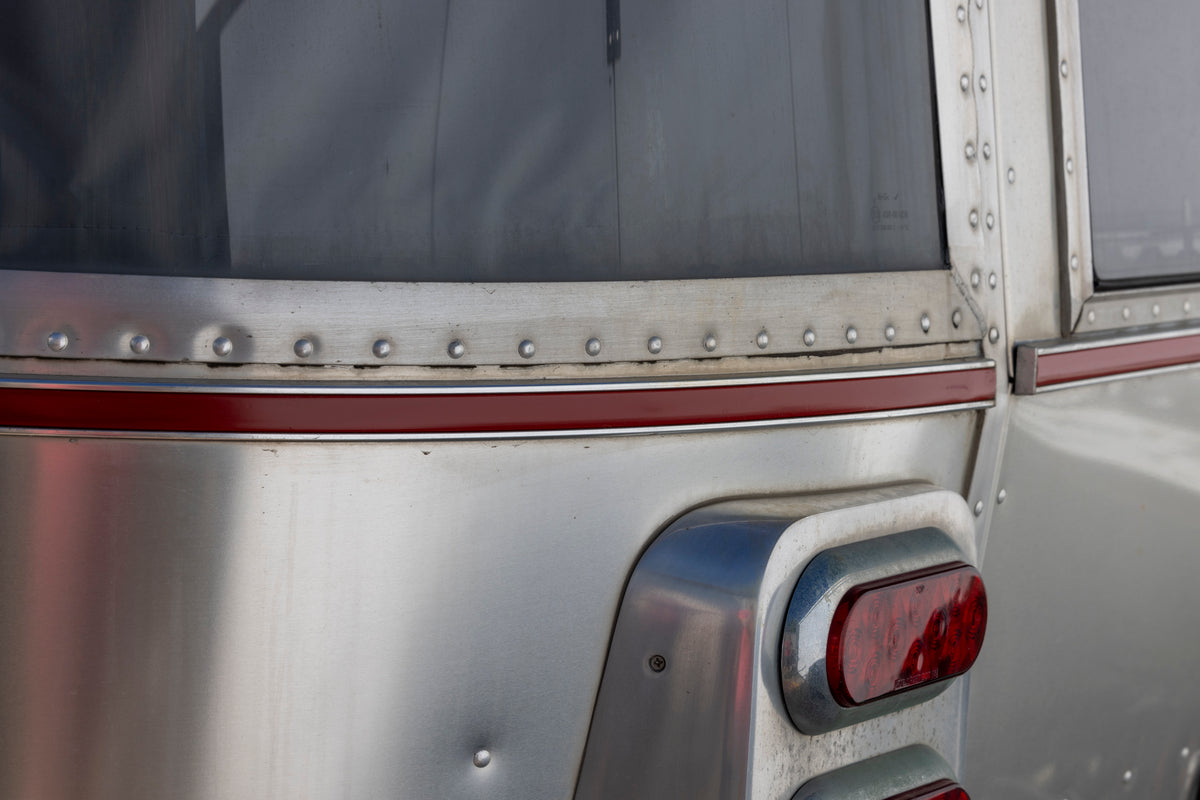 Close-up of the rear corner of a silver metal trailer featuring the Airstream Premium Replacement Belt Trim for Travel Trailers, with a red accent stripe, visible rivets, and a red brake light. Above, a slightly dirty tinted window highlights the sleek lines provided by the aluminum trim.
