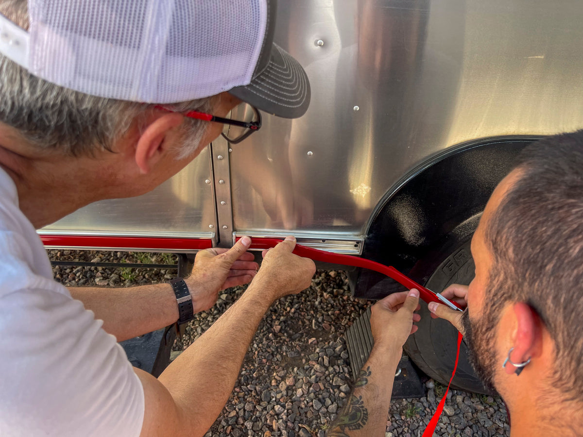 Two people install an Airstream Premium Replacement Rub Rail for Travel Trailers along the lower edge of a shiny, riveted aluminum Airstream near the wheel. One wears a white shirt and hat, while the other has short hair and a tattoo. Gravel covers the ground as they work together to apply the new red rub rail.