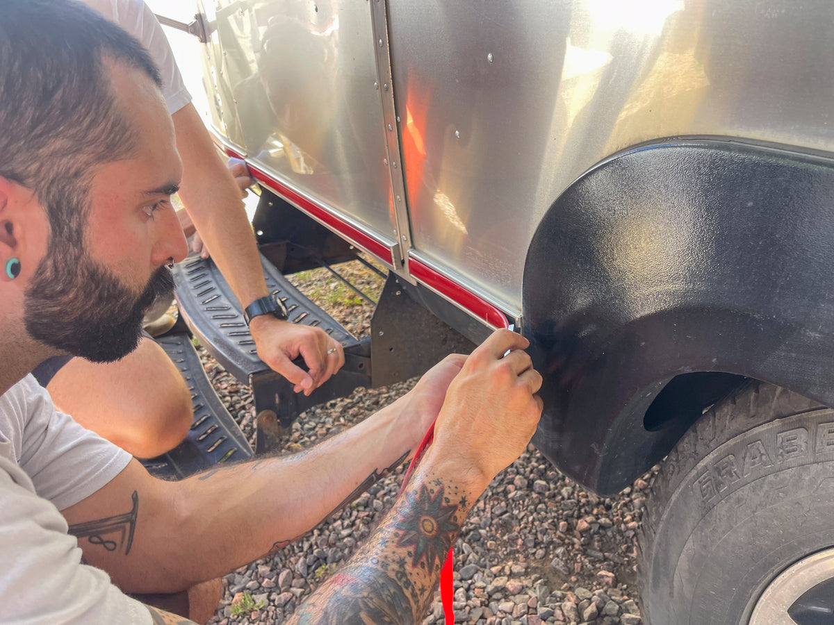 A person with tattoos and gauged earrings carefully installs an Airstream Premium Replacement Rub Rail for Travel Trailers along the edge of a riveted aluminum Airstream, while another person watches and assists nearby. The ground is covered with small gravel.