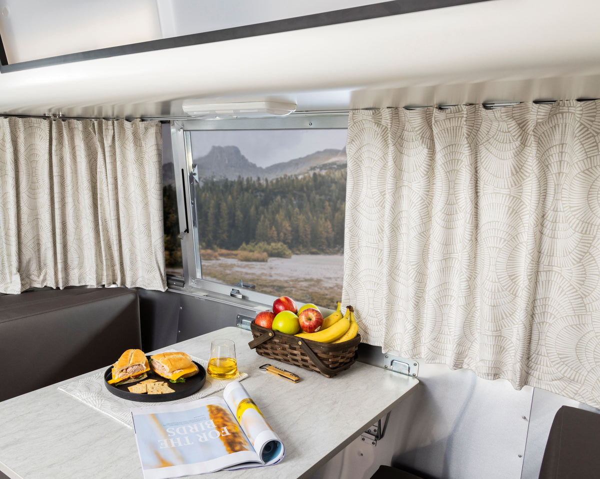 Inside a camper, a table holds a plate with sandwiches, a glass of juice, fruit in a basket, and an open magazine. Airstream Printed Custom Curtains for Bambi Travel Trailers in soft beige frame a window revealing a scenic mountain and forest landscape.