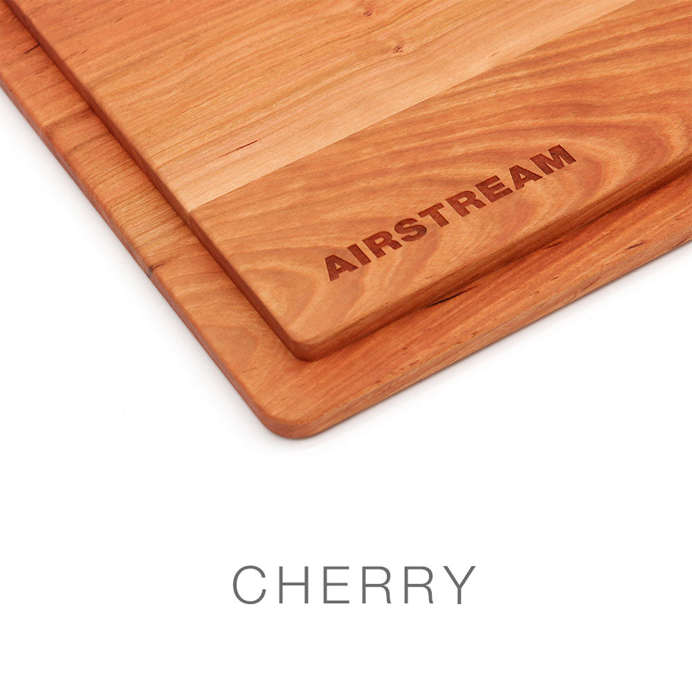 A close-up of a rectangular cherry wood cutting board with AIRSTREAM engraved on it. This item is the Airstream Custom Sink Cutting Board for Basecamp, designed as a premium accessory. The word CHERRY appears below the board against a white background.