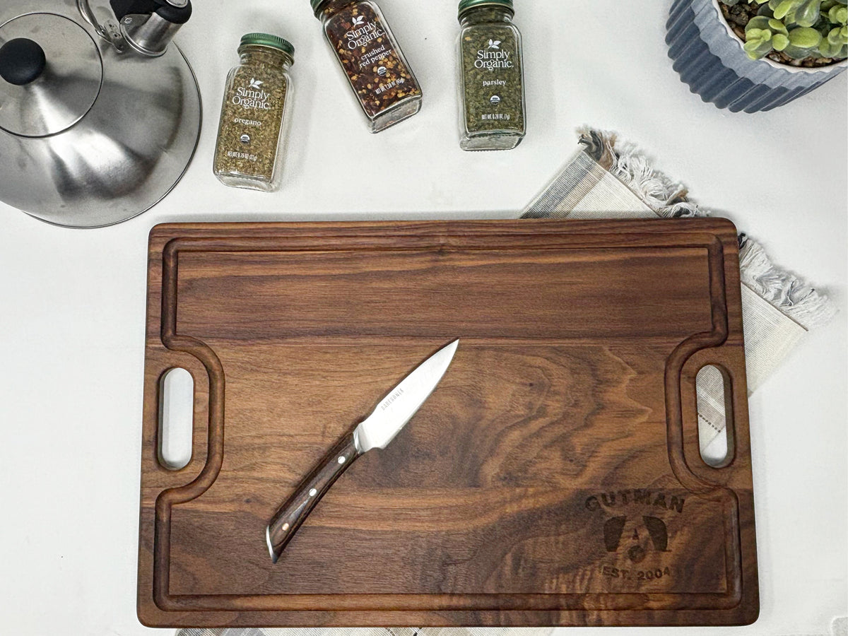 An Airstream Personalized Cutting and Serving Board engraved with “GUTMAN EST. 2004” sits on a white table with a knife resting on top. Nearby, jars of spices, a teapot, a fringed napkin, and a potted plant add to the scene—perfect for those who appreciate custom cutting boards.