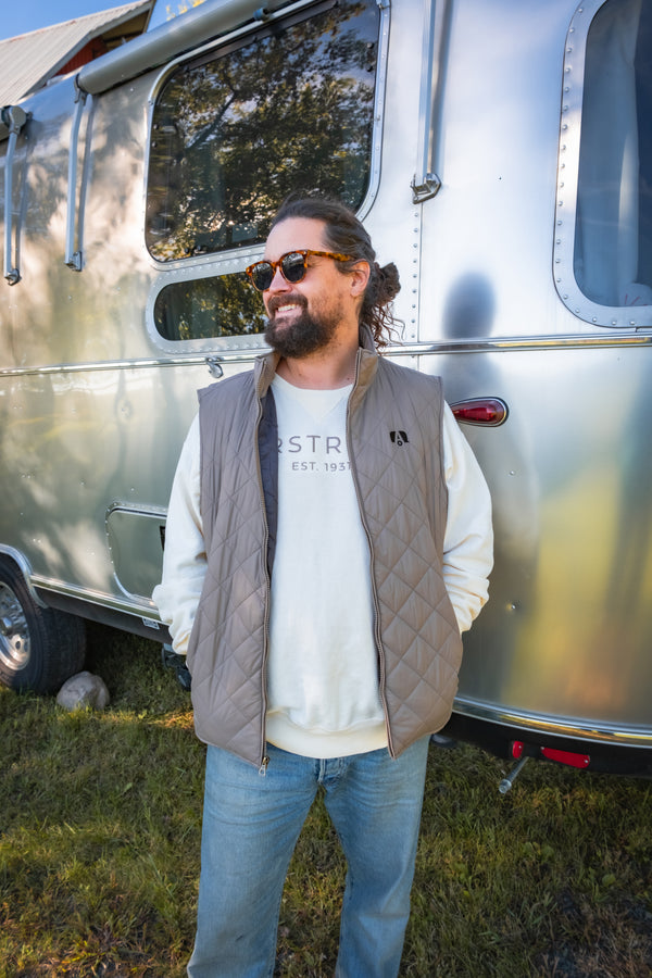 A man with a beard and sunglasses stands smiling in front of a silver Airstream trailer, wearing the Airstream Vintage Diamond Quilted Vest over a light sweatshirt and blue jeans.