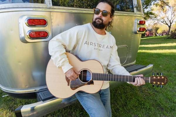 A man with sunglasses and a beard strums an acoustic guitar as he sits on the bumper of a silver Airstream trailer in a grassy outdoor setting, dressed in the Airstream Tonal Comfort Wash Crew Sweatshirt for that perfect touch of vintage style.