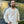 A bearded man with long hair, dressed in jeans and the cream Airstream Tonal Comfort Wash Crew Sweatshirt in a vintage style, stands smiling in front of a gleaming silver Airstream trailer. He holds sunglasses in one hand, with trees reflected behind him outdoors.