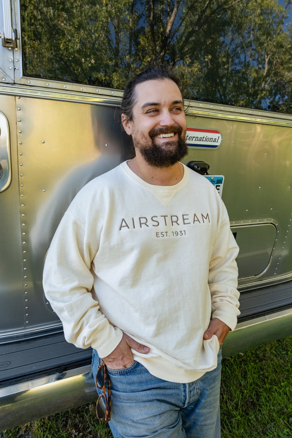 A bearded man with long hair, dressed in jeans and the cream Airstream Tonal Comfort Wash Crew Sweatshirt in a vintage style, stands smiling in front of a gleaming silver Airstream trailer. He holds sunglasses in one hand, with trees reflected behind him outdoors.