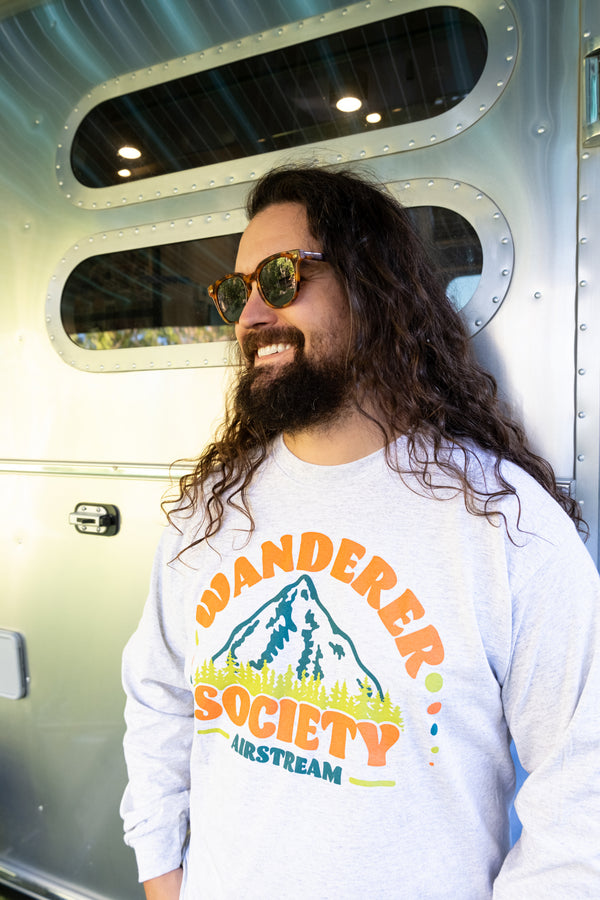 A man with long curly hair and sunglasses smiles while standing in front of an Airstream trailer, wearing a gray Airstream Wanderer Society Long Sleeve T-Shirt featuring a mountain graphic—the perfect choice for any travel t-shirt enthusiast.