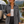 Wearing the Airstream Women’s Long Sleeve Twill Overshirt and black pants, a woman stands by a lakeside cabin and an Airstream trailer, framed by pine trees and snow-capped mountains.