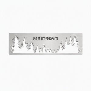 The Airstream Forest Scene Screen Door Guard is a custom-made silver metal plate featuring the word “AIRSTREAM” cut out, with surrounding cut-out silhouettes of pine trees, designed as an accent for Airstream screen doors and set against a white background.