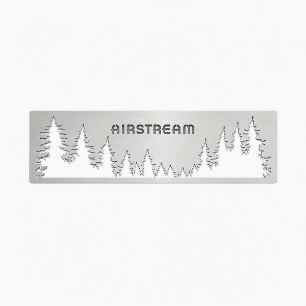 The Airstream Forest Scene Screen Door Guard is a custom-made silver metal plate featuring the word “AIRSTREAM” cut out, with surrounding cut-out silhouettes of pine trees, designed as an accent for Airstream screen doors and set against a white background.