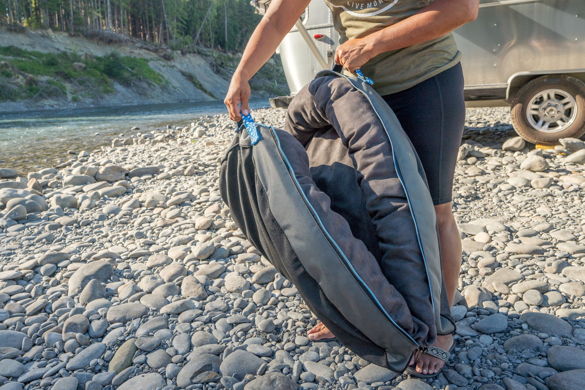 A person stands on a rocky riverbank, holding the Airstream Travel Pet Bed—a large, soft, and foldable portable pet bed with convenient handles. An Airstream trailer and a river in the background highlight this must-have accessory for pets on travel and adventure.