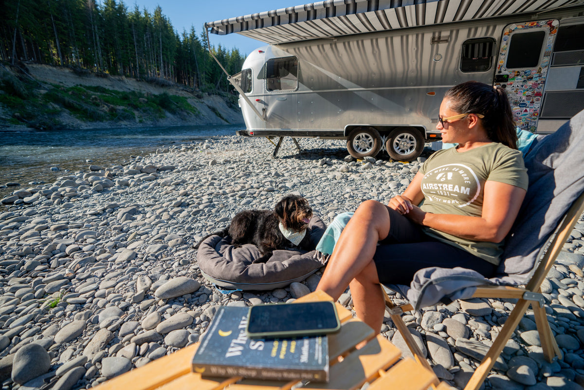 A woman sits in a chair by a river next to an Airstream trailer, smiling at a black dog lying comfortably on an Airstream Travel Pet Bed. A table with books and a phone is in the foreground, and trees are in the background.