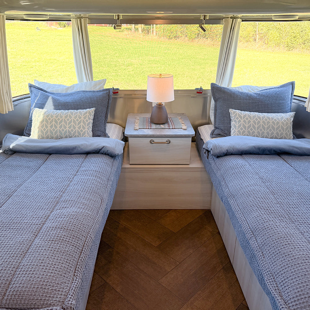 Two twin beds with blue Airstream Custom Fit Dual Comfort Beddy’s Flying Cloud Travel Trailers quilted bedding and patterned accent pillows in a travel trailer, separated by a nightstand with a lamp.