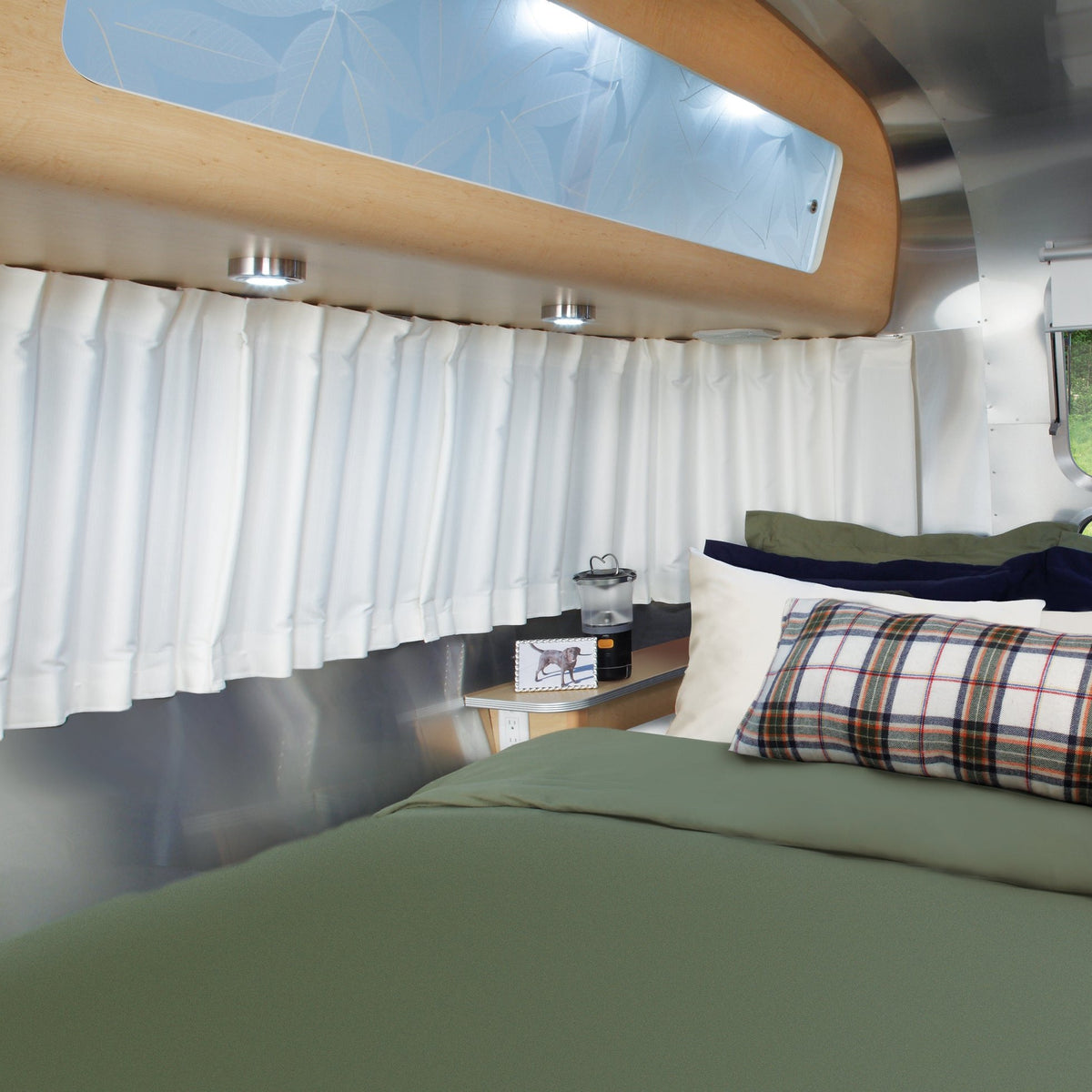 A cozy bed with green bedding and plaid pillows sits beside a small shelf with a lamp, picture frame, and candle in a modern RV interior featuring Airstream Solid Custom Curtains for Eddie Bauer Travel Trailers, white drapes, and warm wood accents.