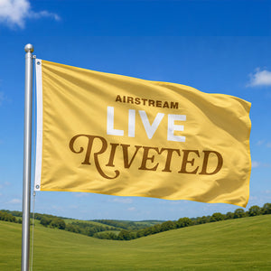 Yellow flag with 'Airstream Live Riveted' text against a blue sky and green landscape