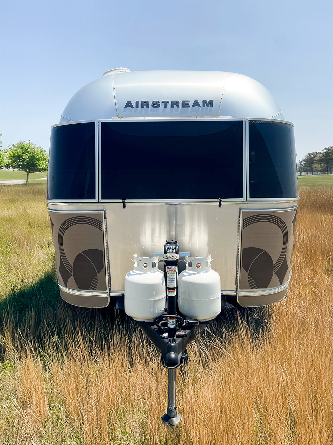 Front view of an Airstream trailer parked in tall, dry grass with two white propane tanks attached to the hitch and a gleaming Airstream Rock Guard from the Rock Wraps Collab Collection protecting the front; a tree and blue sky are visible in the background.