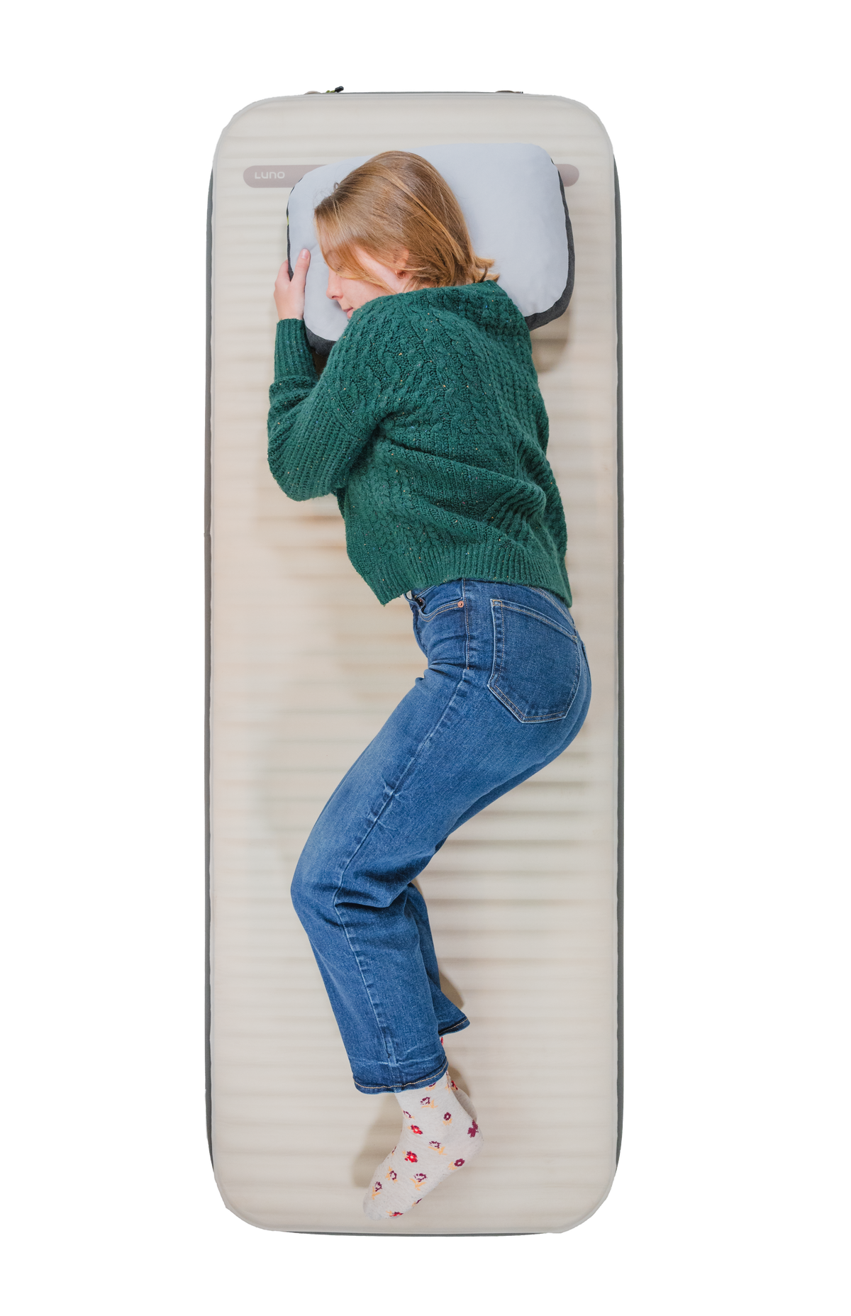 A woman in a green sweater and jeans lies on her side, viewed from above, on the beige Air + Foam Pro Camping Mattress by Luno, using a gray pillow.
