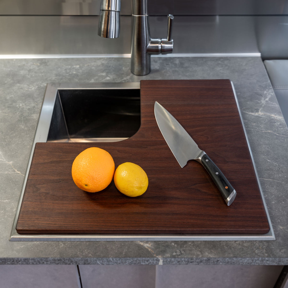 An Airstream Custom Sink Cutting Board for International Travel Trailers, topped with an orange, a lemon, and a chef’s knife, is positioned over a modern kitchen sink featuring a marble countertop and metal faucet.