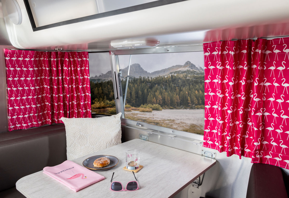 A travel trailer interior featuring a breakfast table, sunglasses, a pillow, a pastry and drink, and Airstream Printed Custom Curtains for Pottery Barn Travel Trailers framing a scenic mountain and forest view through the window.