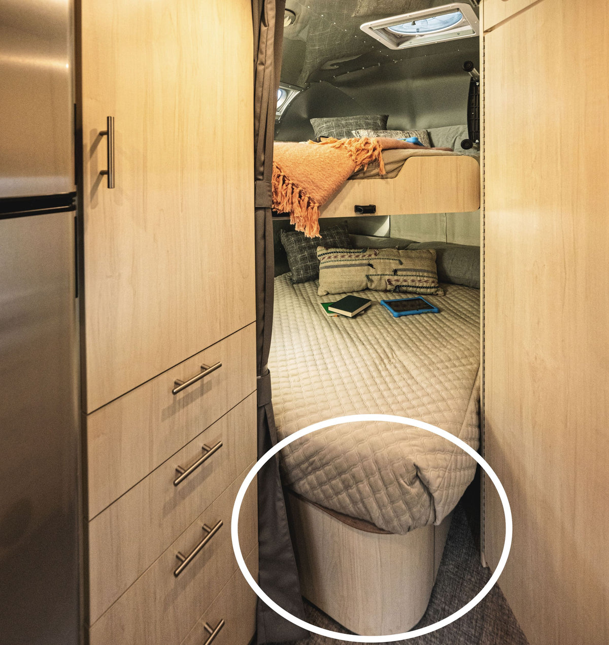 A cozy RV bedroom with a neatly made bed, orange blanket, and several books; a wooden storage compartment under the bed—highlighted with a white circle—features the Airstream x Steele Canvas Twin Bed Storage Bins.